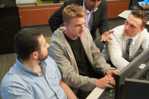 Three students working together around a computer, with a professor behind them making a point while looking at the computer screen
