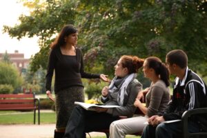 Outside on campus, a professor, standing, discussing a point with three grad students sitting in front of her